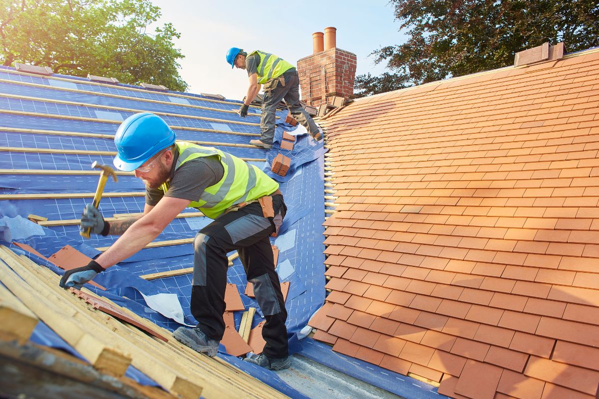 Two men are on a roof, using hammers to work on a construction project, demonstrating teamwork and skill.