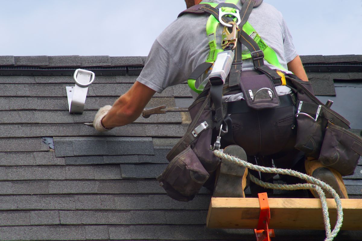 A man climbs a ladder to work on a roof covered with asphalt shingles.